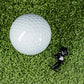 The Blazy Susan Silhouette - Ball Marker by Pins and Aces, a white golf ball, and a black plastic tee rest on green turf—a premium golf accessory set ideal for every enthusiast.