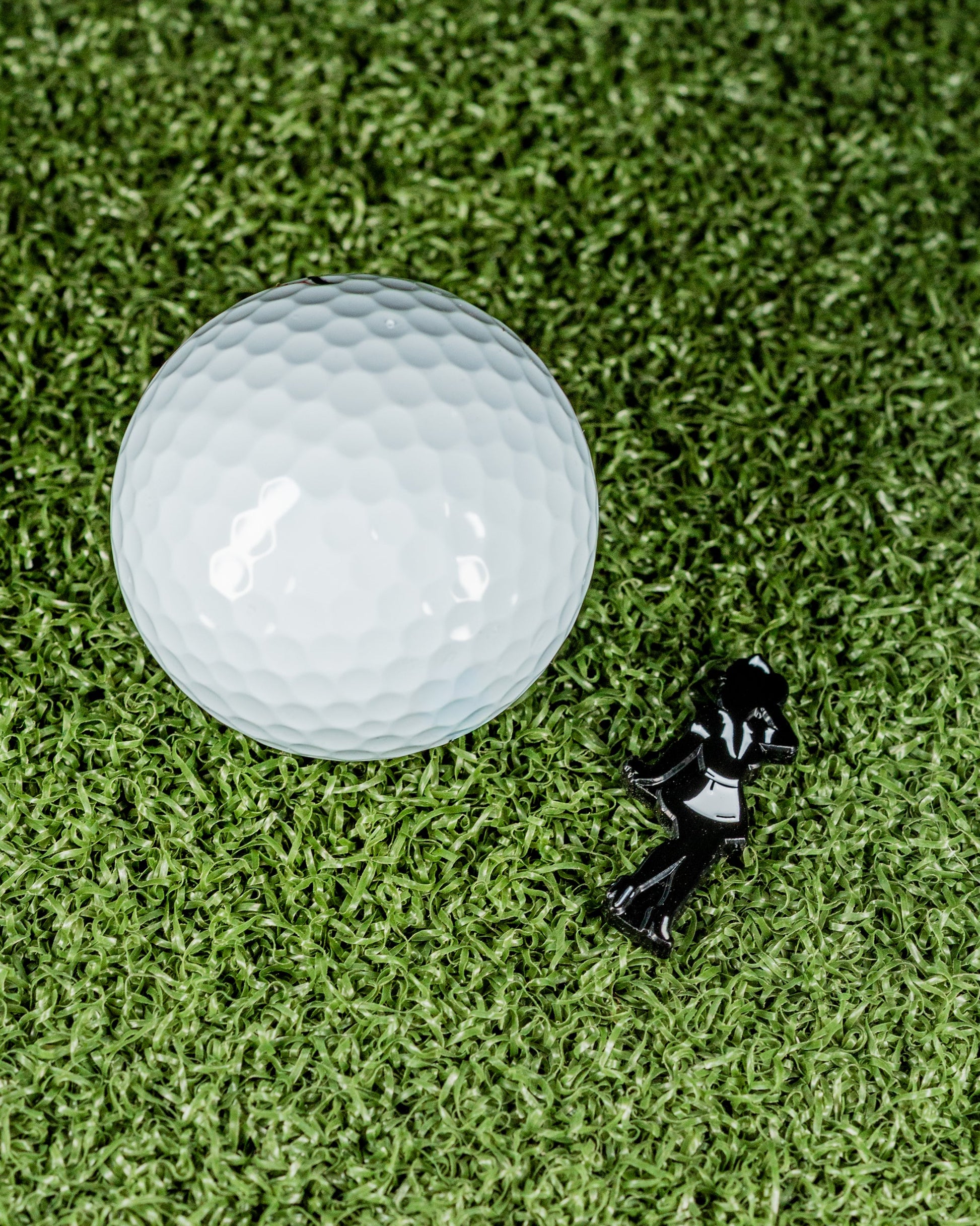 The Blazy Susan Silhouette - Ball Marker by Pins and Aces, a white golf ball, and a black plastic tee rest on green turf—a premium golf accessory set ideal for every enthusiast.