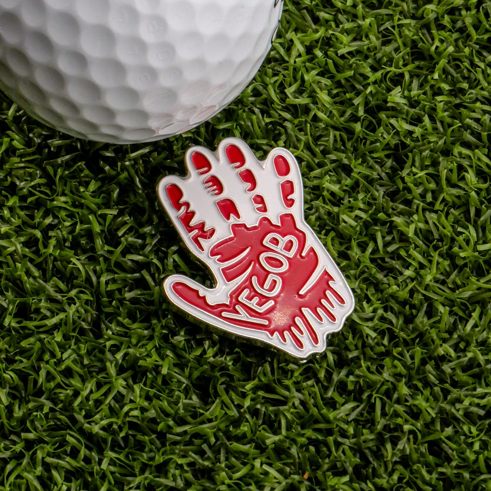The Bloody Bogey RH Ball Marker by Ballmarkers.com, shaped like a red-and-white open hand with "VEGOB," lies on green turf beside a white golf ball, adding a striking touch—like a horror film prop for your next golf round.