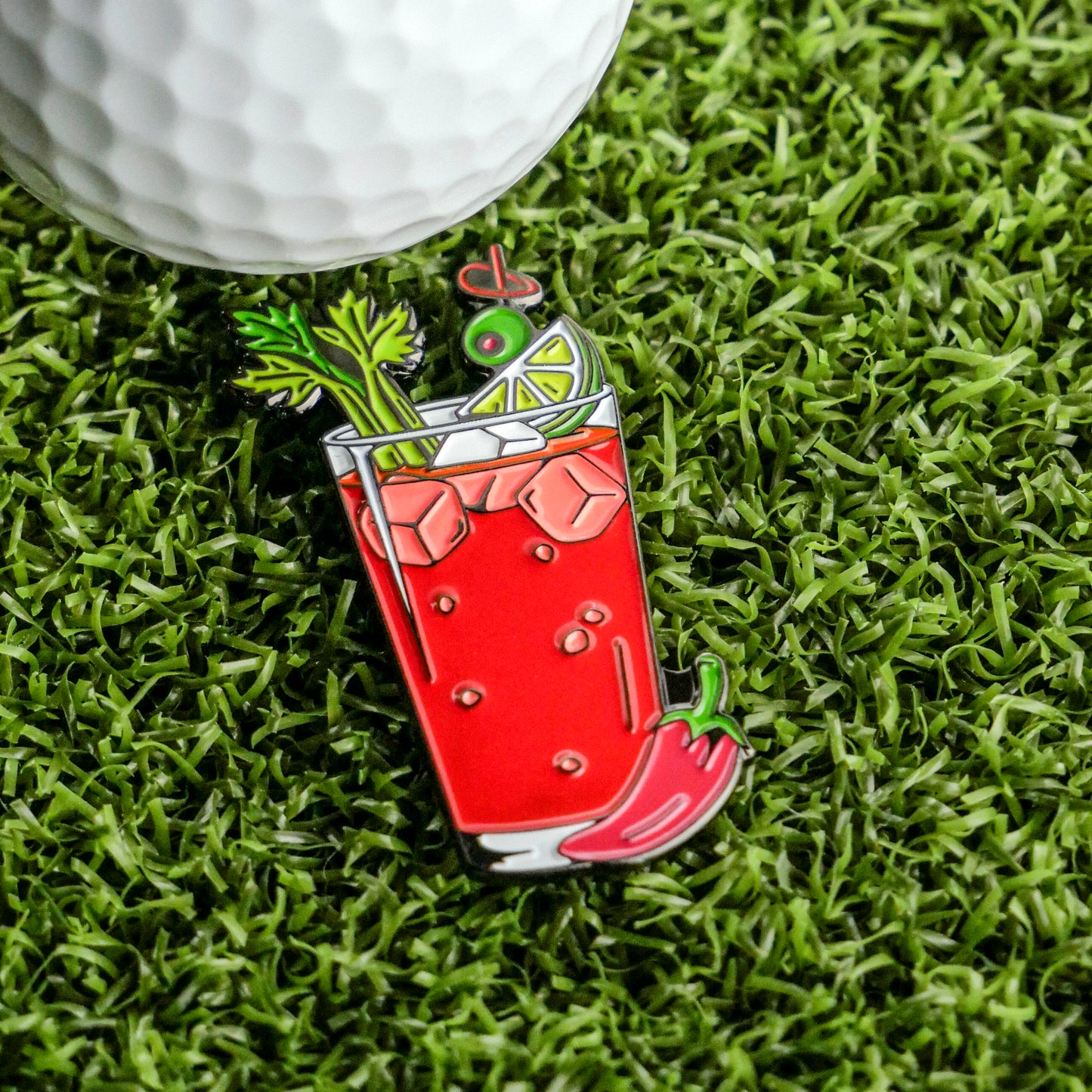 The Bloody Mary Ball Marker by Ballmarkers.com, shaped like the classic cocktail with ice and garnishes, sits on green grass next to a golf ball—a fun brunch golf gift or unique cocktail-themed accessory.