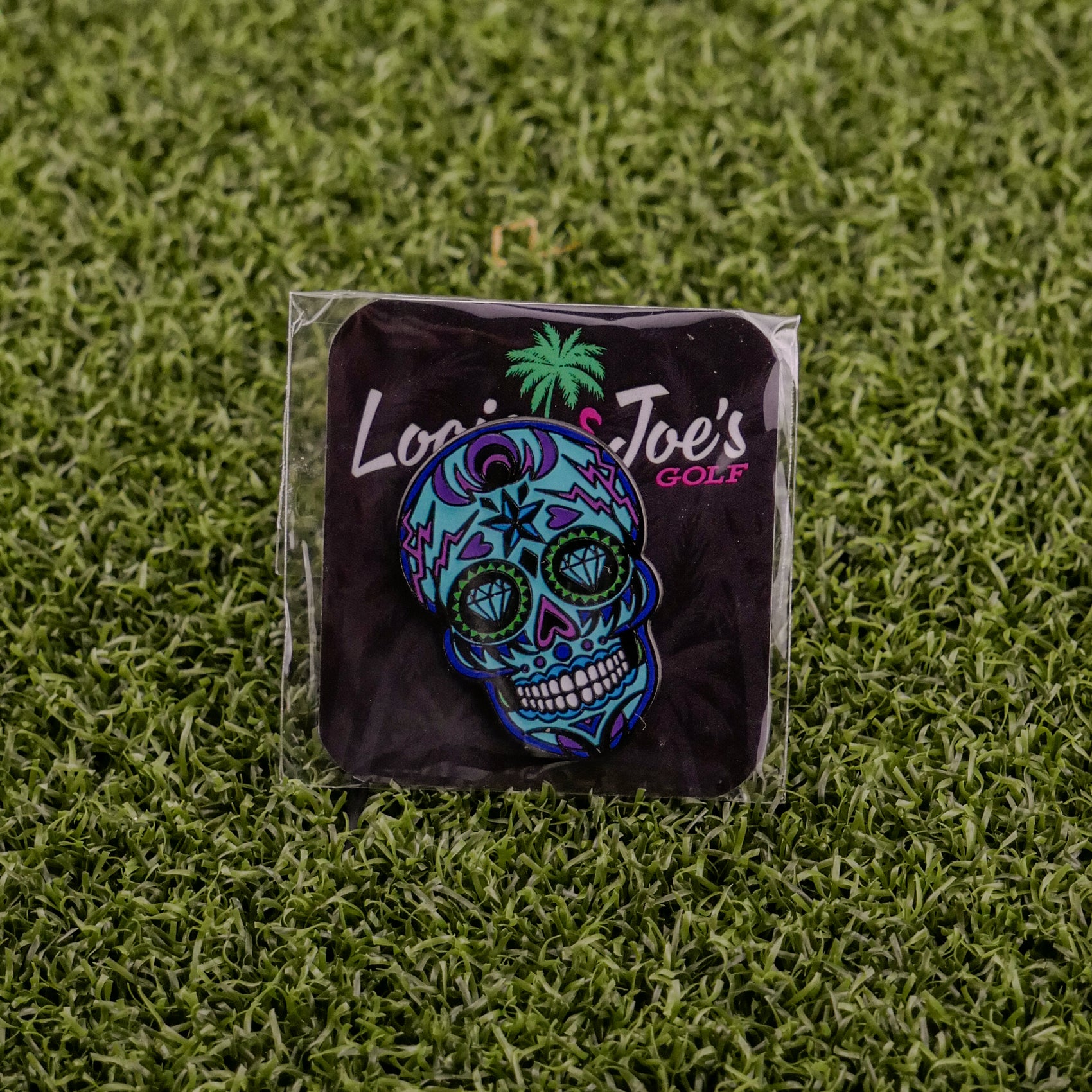 The Blue Sugar Skull ball marker by Logie Joe's Golf features vibrant blue, purple, and green hues on a black card with the palm tree logo—an artistic golf accessory that adds unique flair to your game.
