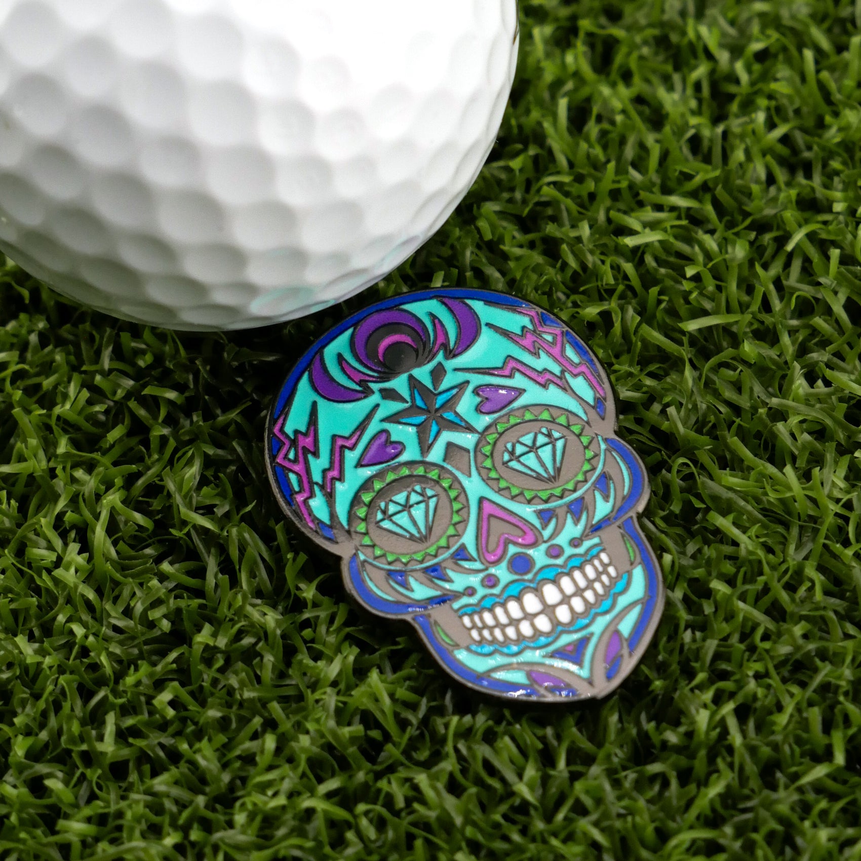 A vibrant Blue Sugar Skull ball marker by Logie Joe's Golf with intricate patterns rests on green grass beside a white golf ball, highlighting stylish, artistic golf accessories.