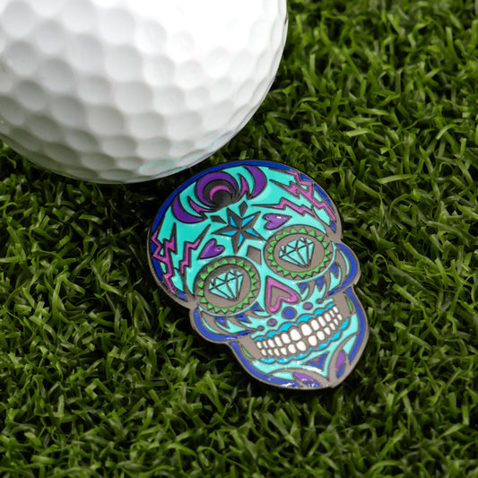The Blue Sugar Skull Ball Marker from Ghouls of the Green by Logie Joe's Golf, featuring turquoise, purple, and green designs, rests on grass beside a white golf ball, highlighting stylish golf accessories.