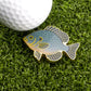 The Logie Joe's Golf Bluegill Ball Marker, featuring gold edges and blue-green scales in a fish shape, sits on artificial grass next to a white golf ball—ideal for those who appreciate premium golf accessories.