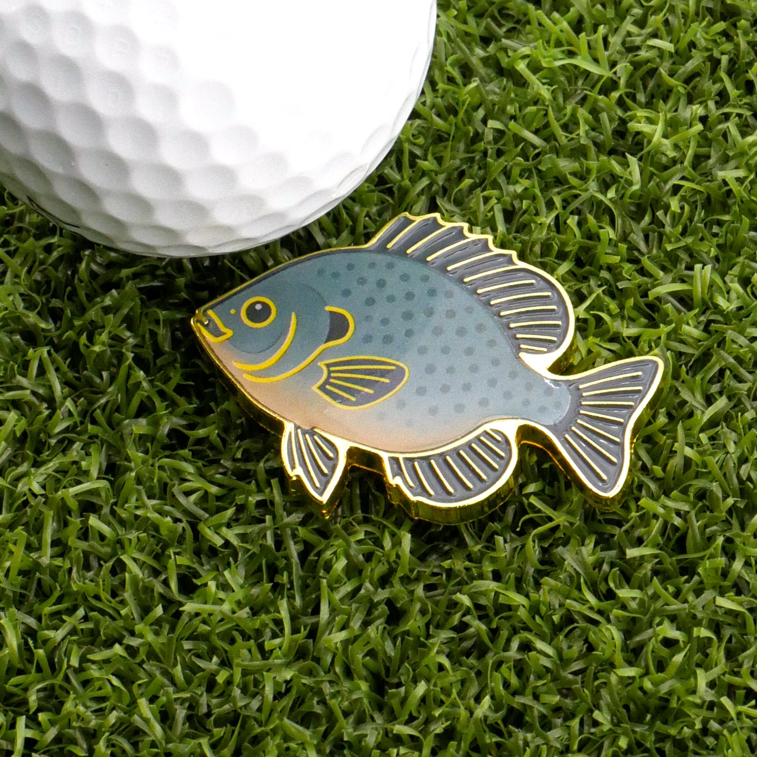 The Logie Joe's Golf Bluegill Ball Marker, featuring gold edges and blue-green scales in a fish shape, sits on artificial grass next to a white golf ball—ideal for those who appreciate premium golf accessories.