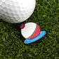 A white golf ball on green grass sits next to Logie Joe's Golf Bobber, a buoy-shaped enamel pin on blue water, used as a unique magnetic ball marker—ideal for those who love premium golf accessories.