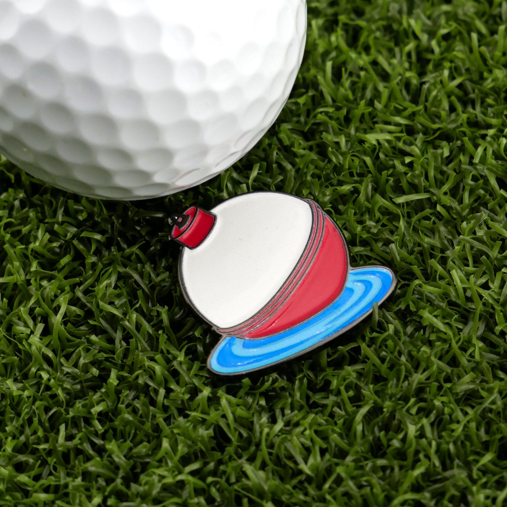 A white golf ball on green grass sits next to Logie Joe's Golf Bobber, a buoy-shaped enamel pin on blue water, used as a unique magnetic ball marker—ideal for those who love premium golf accessories.