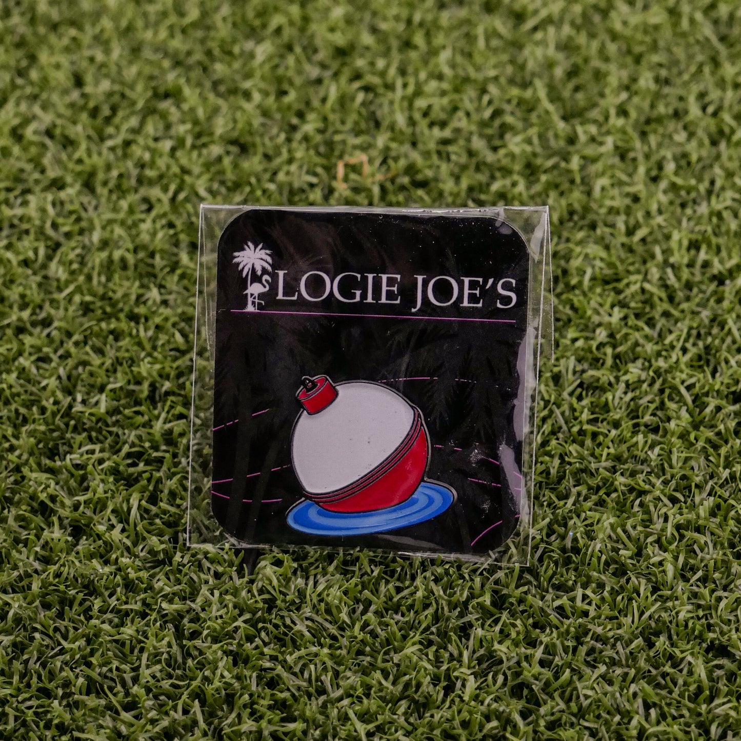 The Bobber by Logie Joe’s Golf is a square pin with a red and white fishing bobber on water, topped by the brand’s palm tree logo. Placed on green turf, it also doubles as a unique magnetic golf ball marker—perfect for premium collections.