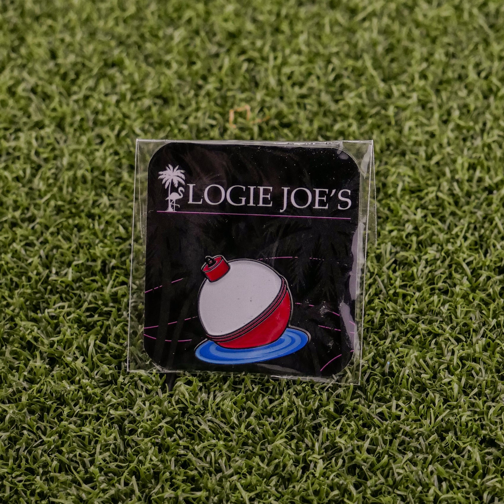 The Bobber by Logie Joe’s Golf is a square pin with a red and white fishing bobber on water, topped by the brand’s palm tree logo. Placed on green turf, it also doubles as a unique magnetic golf ball marker—perfect for premium collections.