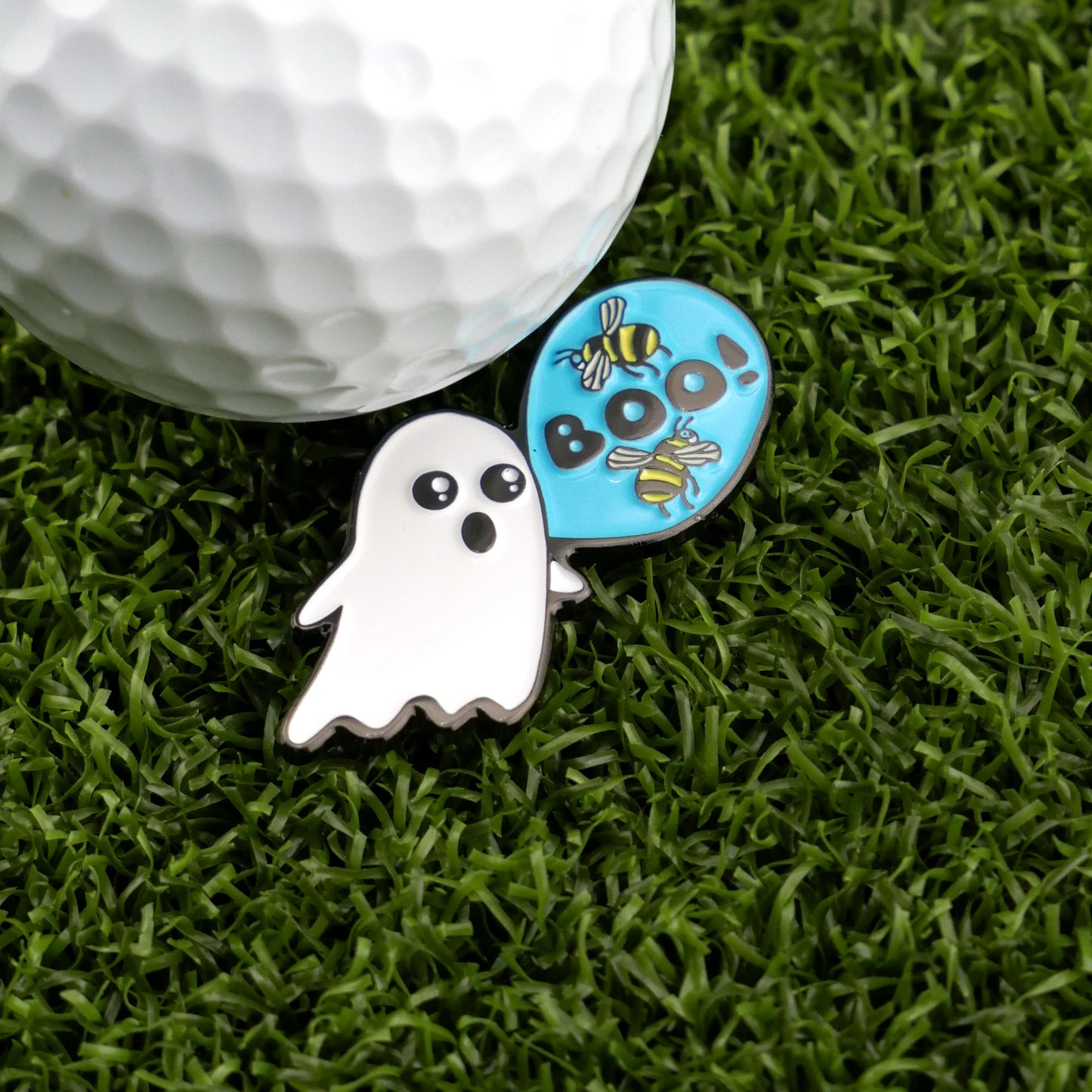 A Logie Joe's Golf "Boo Bees" ball sits on grass next to ghost-shaped markers and a blue “Boo!” pin with bees—ideal for fans of fun golf accessories.