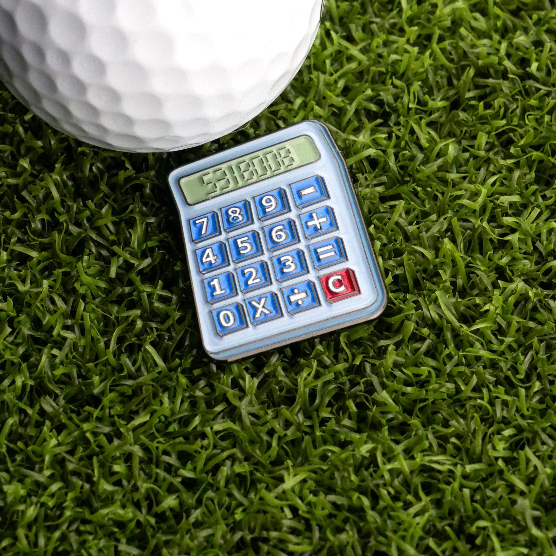 The Boobies Calculator Ball Marker by Ballmarkers.com, a blue toy calculator with white and red buttons, rests on green artificial grass next to a large white golf ball—perfect as a unique golf accessory.