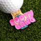 The Bang Bang Booty Gang Ball Marker by Logie Joe’s Golf is a metal pin with “BOOTY GANG” in bold pink letters and a cartoon jeans design, shown on green turf next to a golf ball.