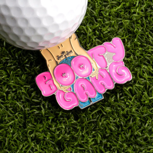 The Bang Bang Booty Gang Ball Marker by Logie Joe’s Golf is a metal pin with “BOOTY GANG” in bold pink letters and a cartoon jeans design, shown on green turf next to a golf ball.