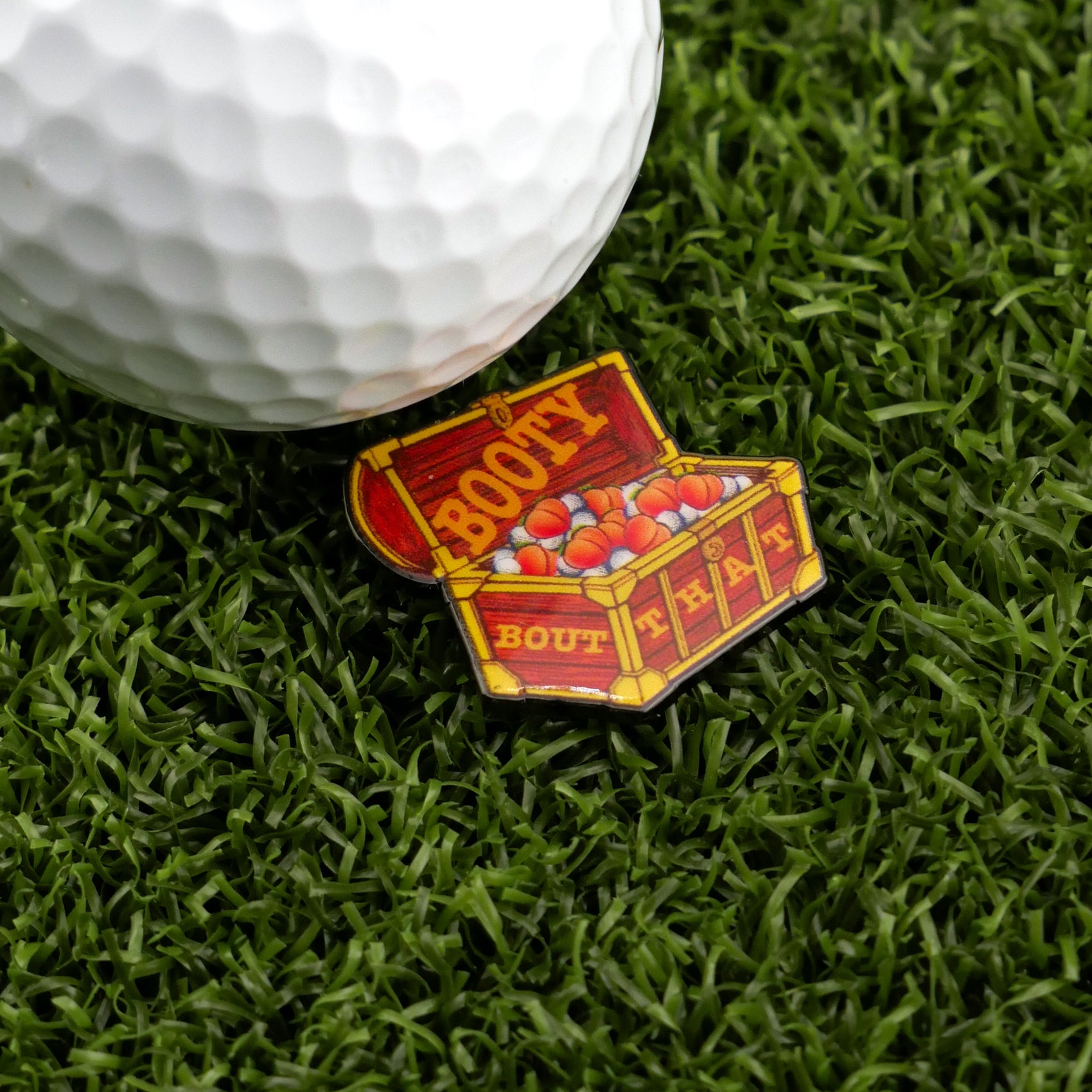 A close-up of a golf ball on artificial grass beside Logie Joe’s Golf "Bout That Booty," a colorful treasure chest–shaped ball marker.