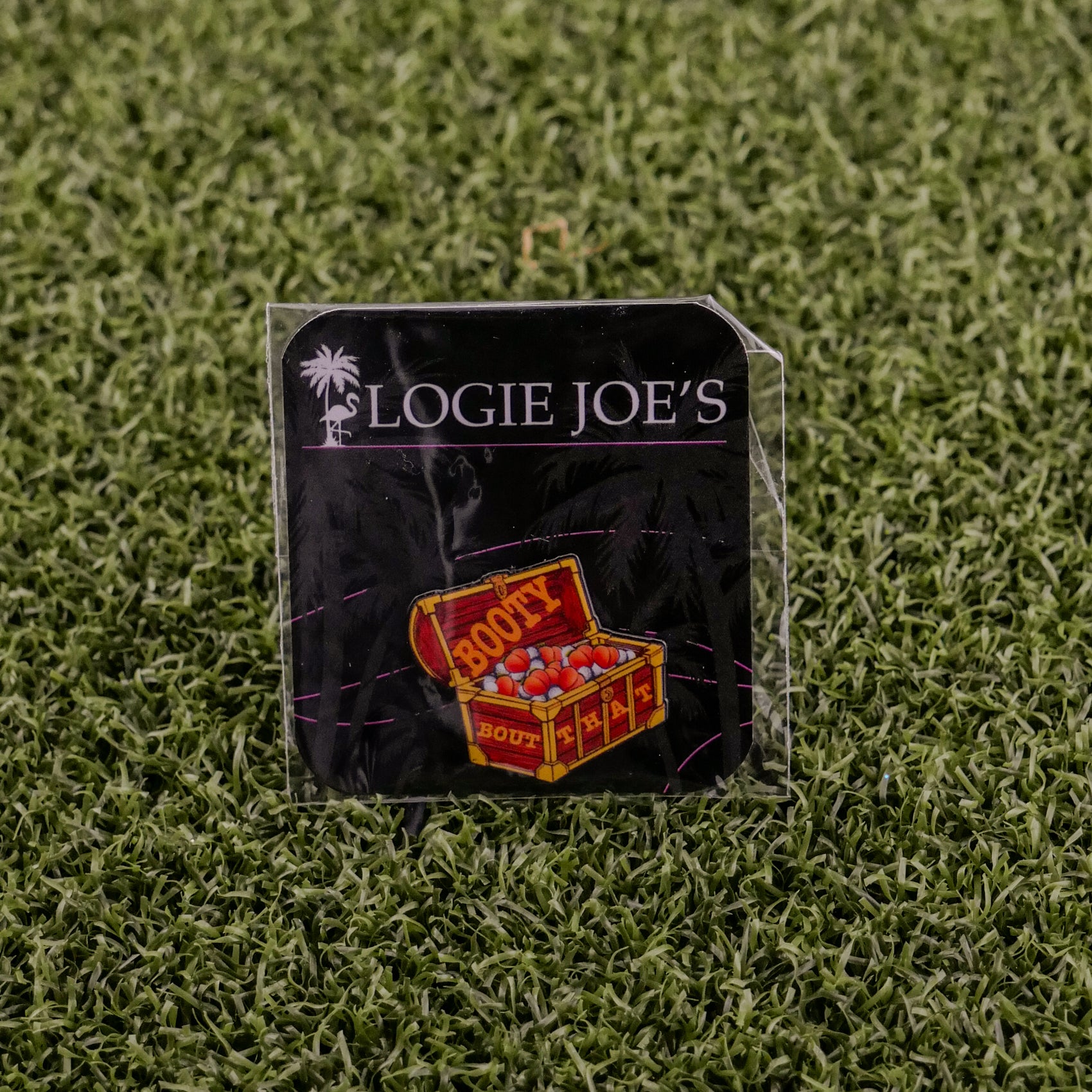 The "Bout That Booty" pin from Logie Joe's Golf features a colorful treasure chest and doubles as a premium ball marker, packaged on branded carding with an artificial grass background.