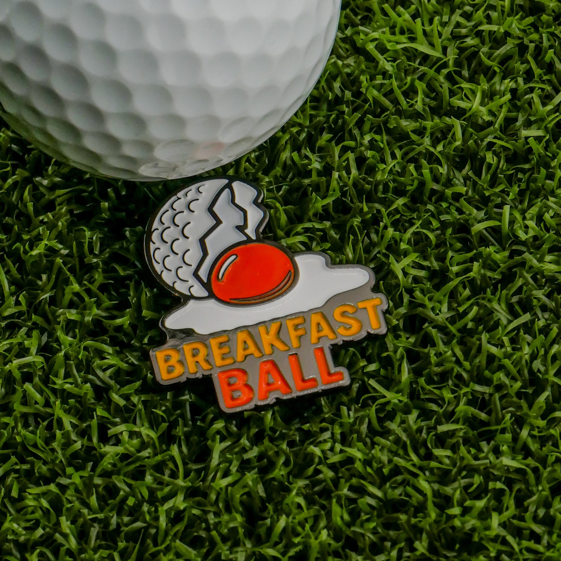 The Breakfast Ball (Sunnyside Up) Ball Marker by Logie Joe’s Golf features a cracked golf ball as a fried egg and “BREAKFAST BALL” below in yellow and orange—a quirky choice for fans of unique personalized golf accessories.