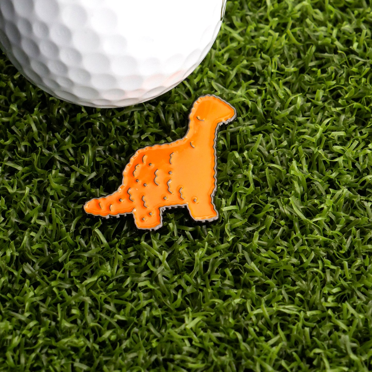 A bright orange Brontosaurus Dinosaur Nugget Ball Marker by Ballmarkers.com rests on green artificial grass beside a white golf ball.