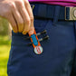 A person wearing navy blue pants clips THE BYRD GOLF CLYP - White Swan by The Byrd, LLC—with free tees and a ball marker—to their pocket. Their hand is near the clip, and they wear a belt with a decorative buckle.