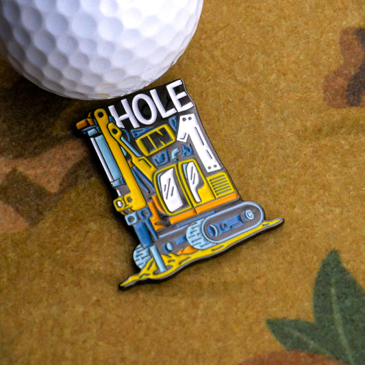 A close-up of the Ace Maker Ball Marker by Ballmarkers.com, a vibrant enamel pin shaped like a yellow excavator with "HOLE" in bold white letters, posed next to a golf ball on a textured brown surface to celebrate excavator golf.
