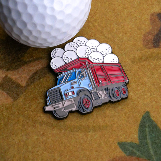 The Ball Hauler Ball Marker from Ballmarkers.com, a decorative pin shaped like a dump truck with lost golf balls, sits on a textured surface beside a golf ball.