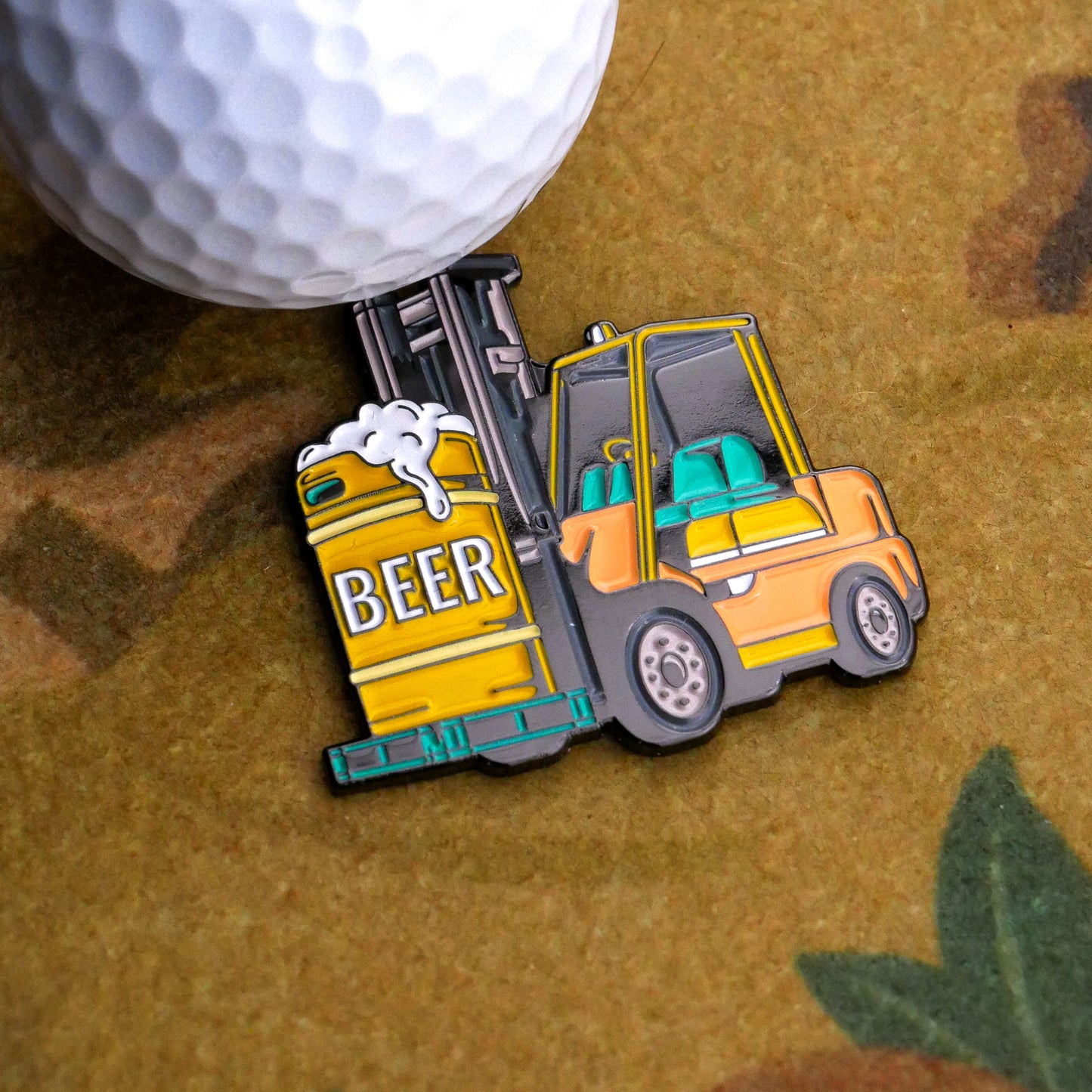 Next to a golf ball is the Ballmarkers.com Beer Delivery Ball Marker, a vibrant enamel pin of a yellow forklift lifting a BEER-labeled keg—ideal for golfers who love beer and camaraderie on the course.