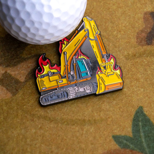 The Burnt Out Digger Ball Marker by Ballmarkers.com features a yellow excavator with red flames, shown next to a white golf ball on a textured brown surface.