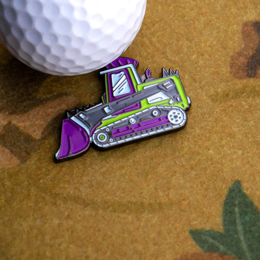 A white golf ball sits beside the Ballmarkers.com Danger Dozer Ball Marker, shaped like a bulldozer with purple and green accents, on a brown surface with a green leaf in the corner—ready for action on the fairway.