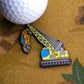 A golf ball sits next to Ballmarkers.com's Heavy Lifter Ball Marker, shaped like a crane hoisting a yellow car, on a brown surface.