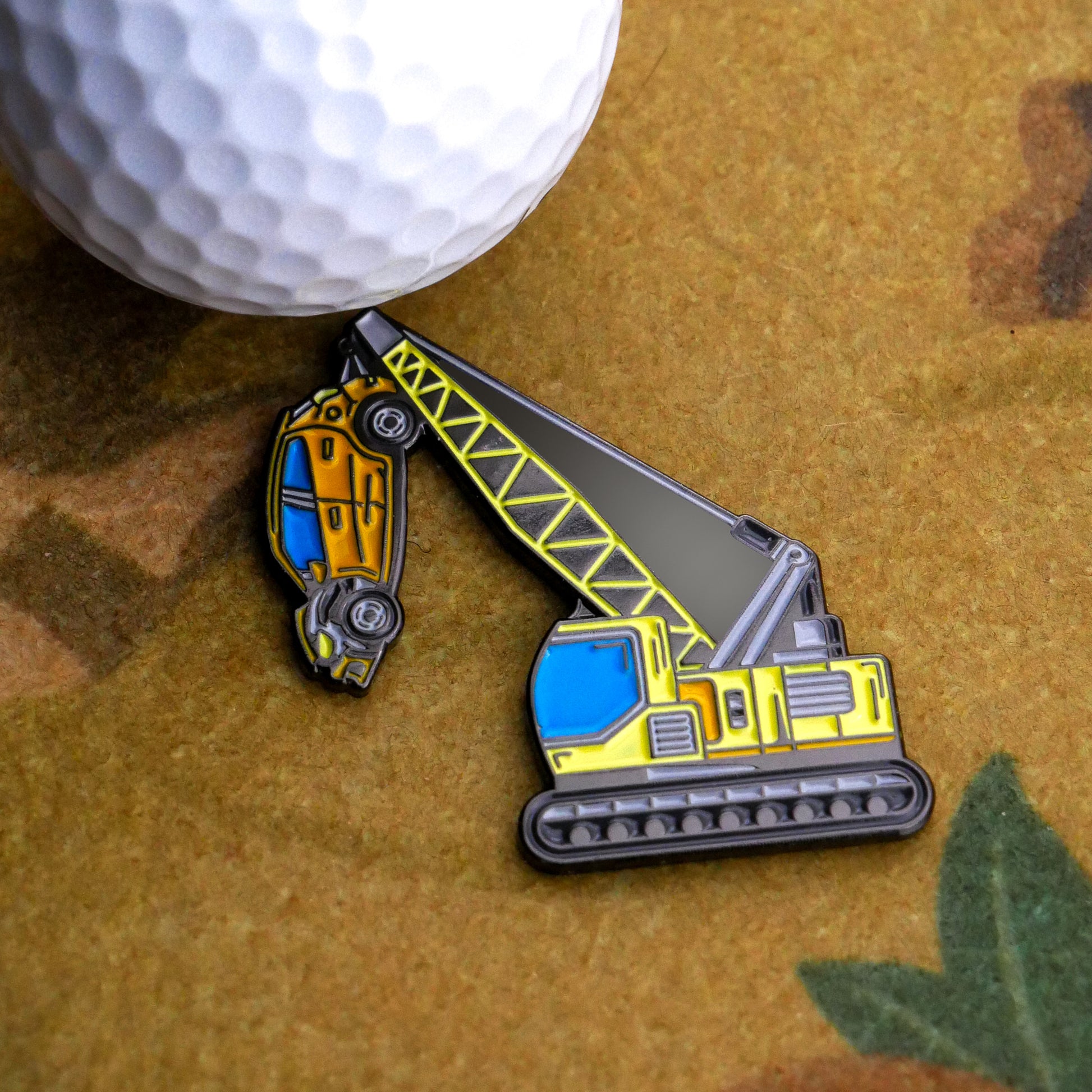 A golf ball sits next to Ballmarkers.com's Heavy Lifter Ball Marker, shaped like a crane hoisting a yellow car, on a brown surface.