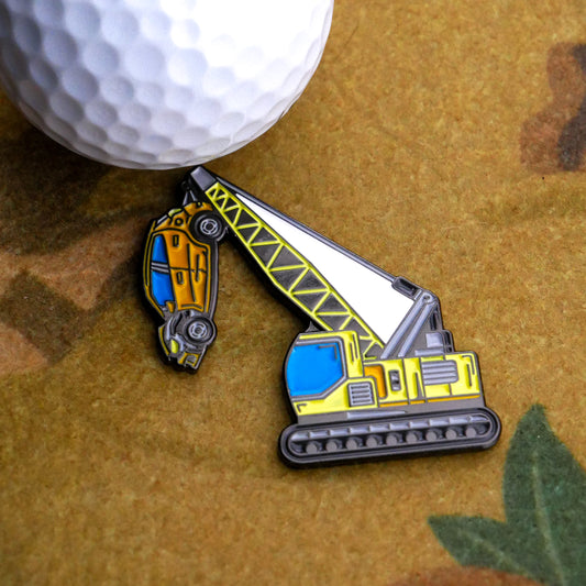 A close-up of a golf ball next to the Ballmarkers.com Heavy Lifter Ball Marker, designed as a crane lifting a yellow car, displayed on a brown patterned surface.