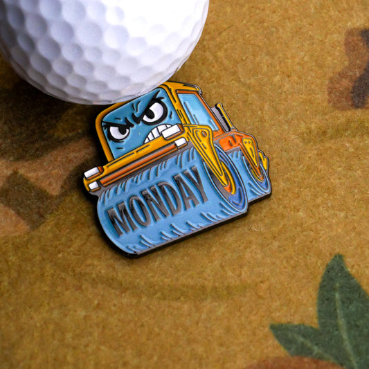 The Monday Steamroller Ball Marker by Ballmarkers.com features a cartoon steamroller with an angry face and "MONDAY" on its blade, ready to flatten your mood on the golf course. Shown next to a white golf ball on a brown surface.