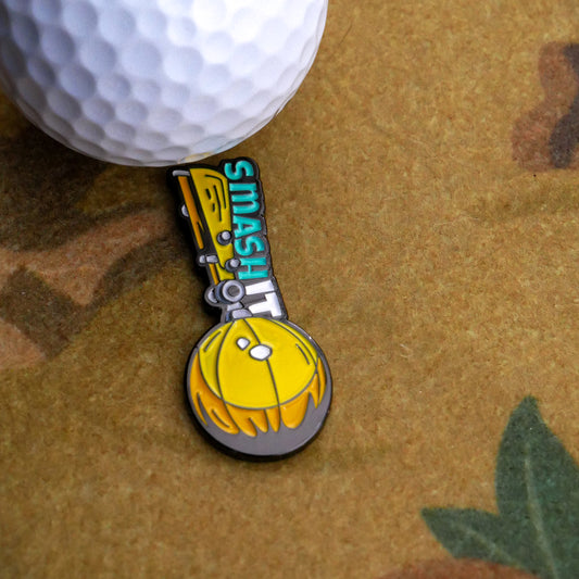 A close-up of a golf ball and club beside Ballmarkers.com's Smash It Ball Marker, an enamel pin with "SMASH IT" text, a yellow helmet, and gray ball on a tan and green patterned surface.