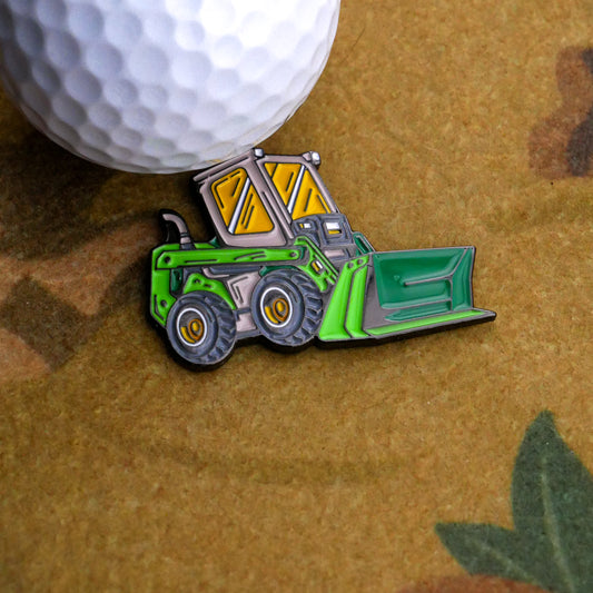 The Un-Loader Ball Marker by Ballmarkers.com, a small pin shaped like a green and yellow skid steer, rests on a brown surface beside a white golf ball.