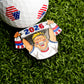 A golf ball with American flag designs rests on grass beside the Ballmarkers.com 2026 Fan Ball Marker—shaped like a cheering fan with a “2026” scarf—making it ideal for soccer fans and collectors alike.