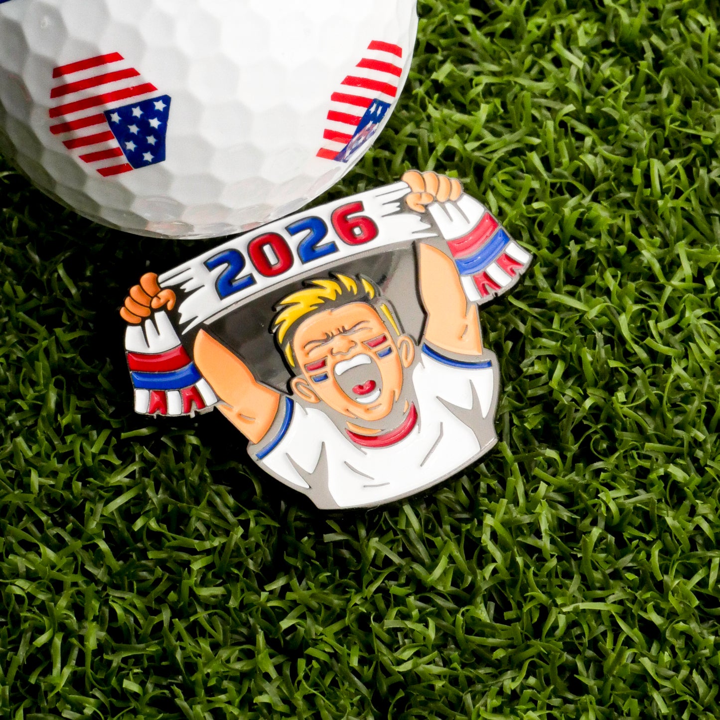 A golf ball with American flag designs rests on grass beside the Ballmarkers.com 2026 Fan Ball Marker—shaped like a cheering fan with a “2026” scarf—making it ideal for soccer fans and collectors alike.