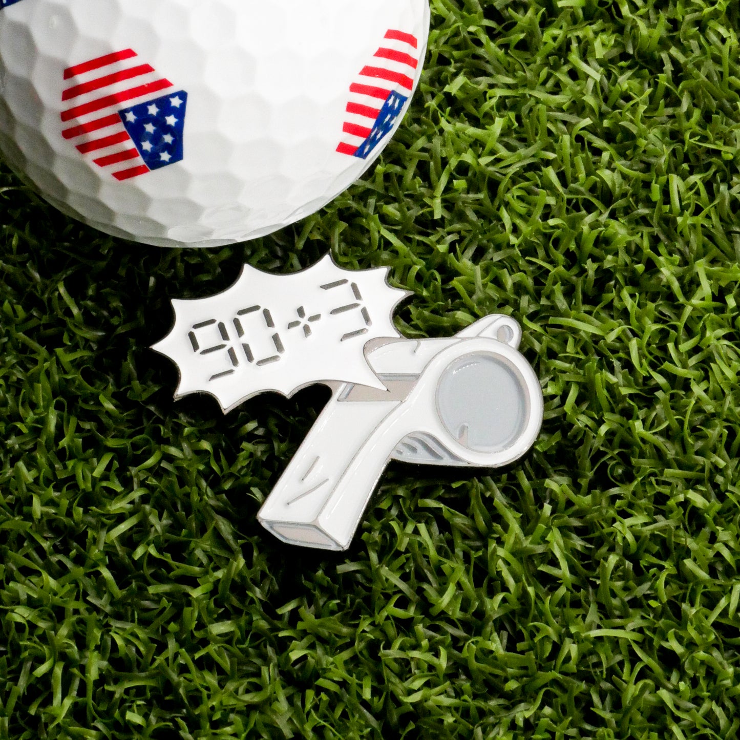 The Extra Time Ball Marker by Ballmarkers.com features an American flag golf ball on grass beside a metallic soccer whistle pin with "90+3" in a comic-style bubble, serving as a unique golf ball marker.