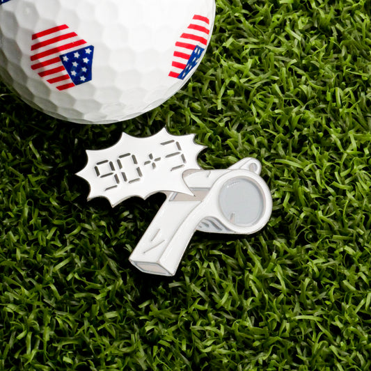 The Extra Time Ball Marker by Ballmarkers.com features an American flag golf ball on grass beside a metallic soccer whistle pin with "90+3" in a comic-style bubble, serving as a unique golf ball marker.