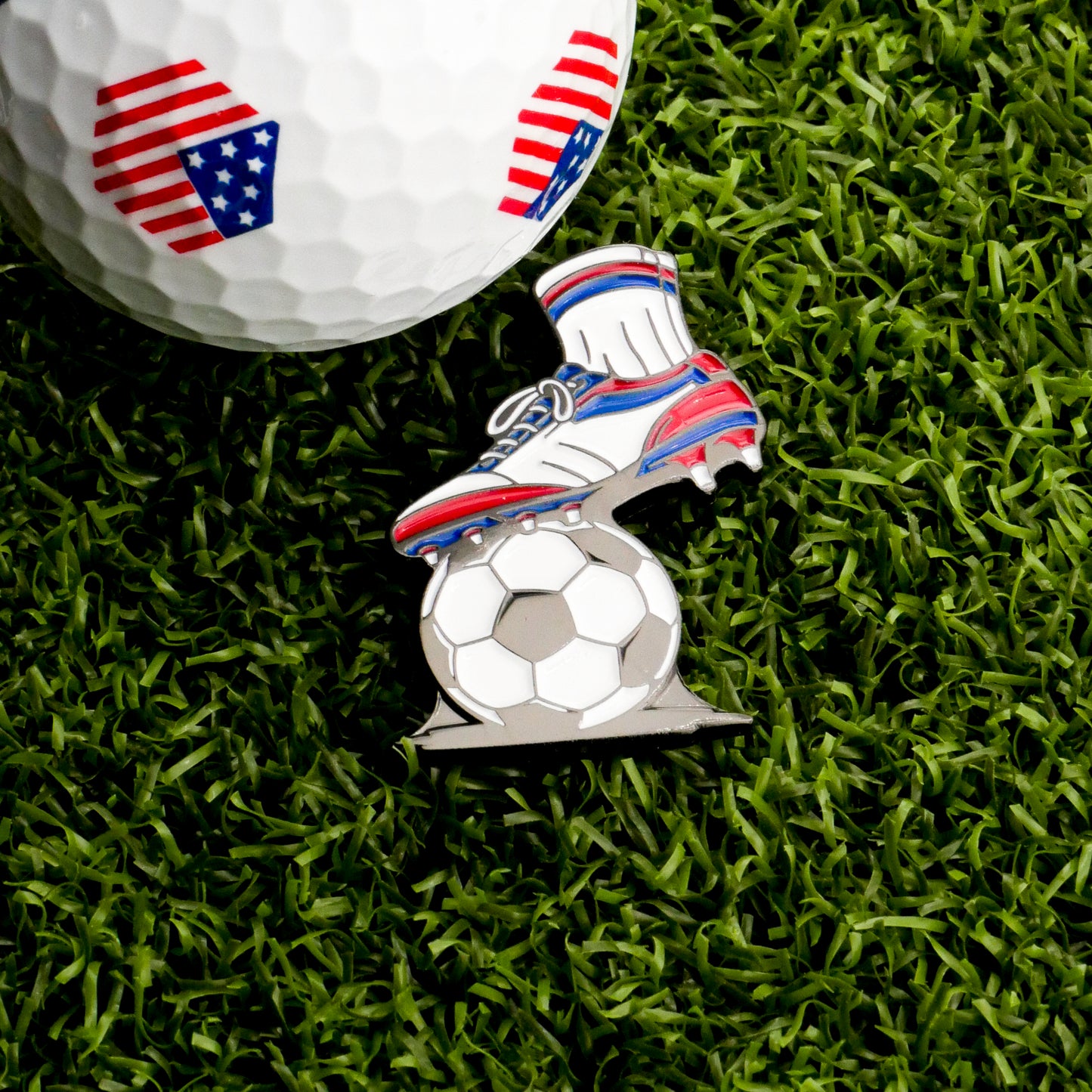 The First Touch Ball Marker by Ballmarkers.com features American flag patterns and includes a pin shaped like a soccer boot kicking a ball, all displayed on green artificial grass.