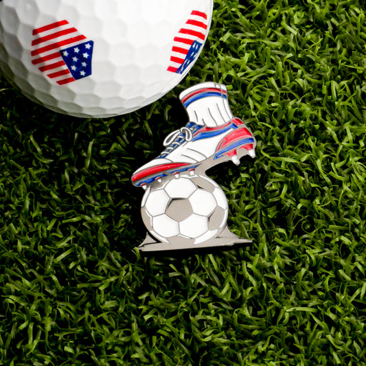The First Touch Ball Marker by Ballmarkers.com features American flag patterns and includes a pin shaped like a soccer boot kicking a ball, all displayed on green artificial grass.