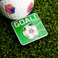 A golf ball with American flag patterns sits on grass beside the Goal! Ball Marker by Ballmarkers.com—a green square accessory with a soccer ball hitting a net and "GOAL!" in bold white letters.