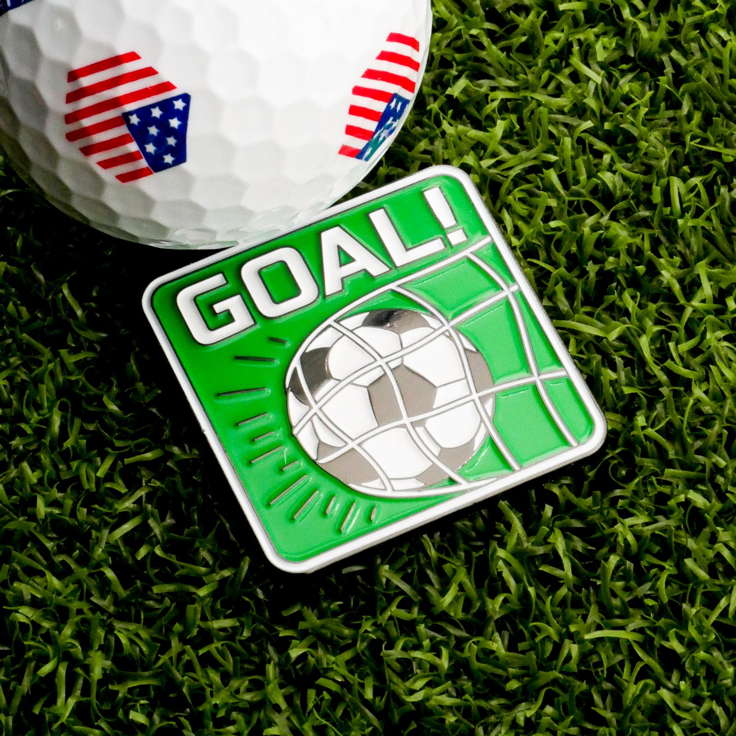 A golf ball with American flag patterns sits on grass beside the Goal! Ball Marker by Ballmarkers.com—a green square accessory with a soccer ball hitting a net and "GOAL!" in bold white letters.