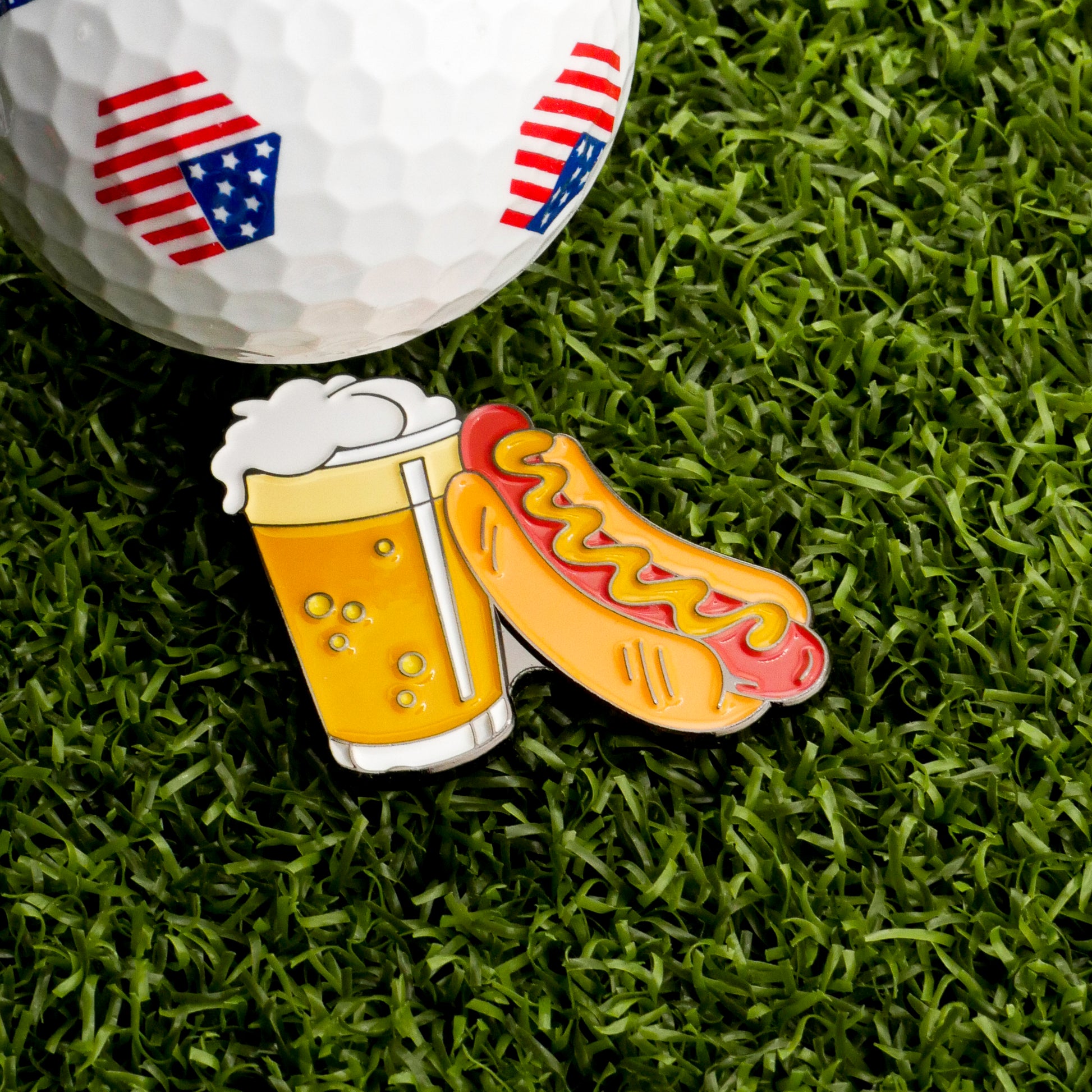 A golf ball with American flag designs sits on grass next to the Ballmarkers.com Glizzy and Beer Ball Marker, which is shaped like a beer mug and a mustard-topped hotdog.