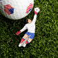 A golf ball with an American flag design sits on grass next to the Goal Tender Ball Marker from Ballmarkers.com—an enamel pin showing a goalkeeper in a white jersey, blue shorts, and red gloves leaping for a save.