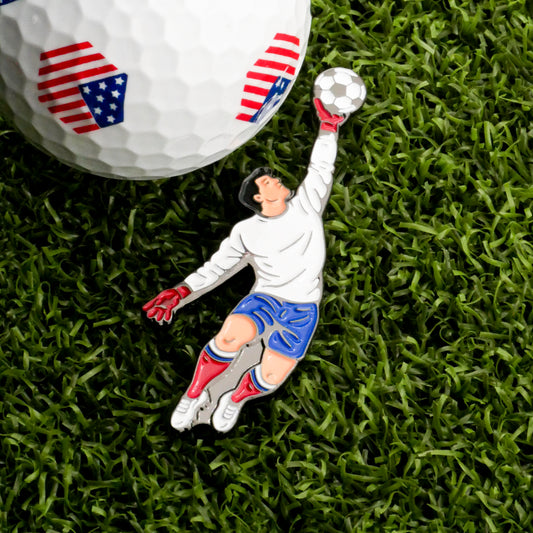 A golf ball with an American flag design sits on grass next to the Goal Tender Ball Marker from Ballmarkers.com—an enamel pin showing a goalkeeper in a white jersey, blue shorts, and red gloves leaping for a save.