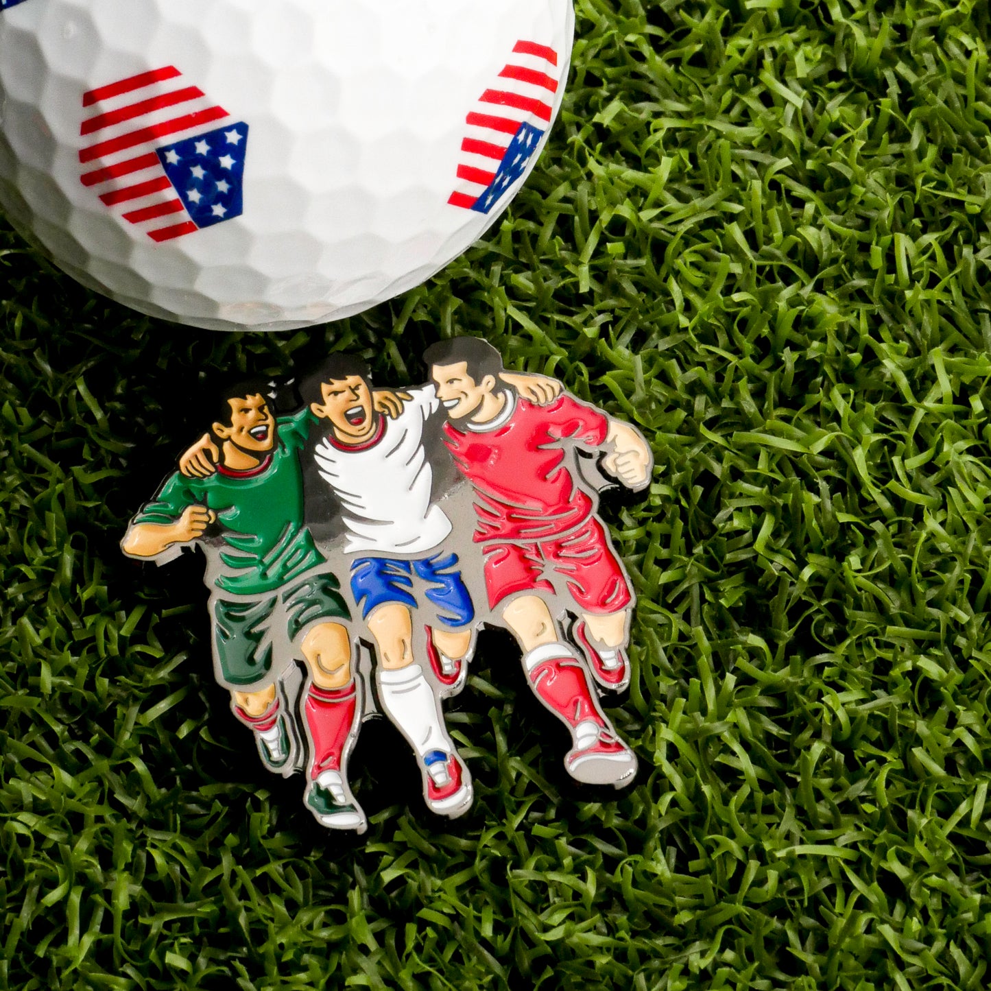 The Host Countries Ball Marker by Ballmarkers.com, featuring American flag patterns, rests on grass beside a soccer enamel pin with three players in green, white, and red uniforms walking arm-in-arm.