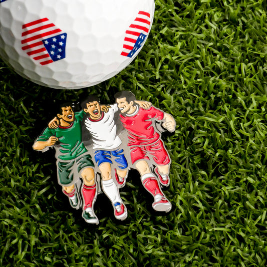 The Host Countries Ball Marker by Ballmarkers.com, featuring American flag patterns, rests on grass beside a soccer enamel pin with three players in green, white, and red uniforms walking arm-in-arm.