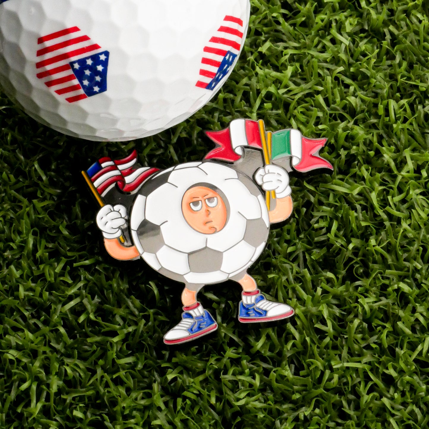 A cartoon soccer ball character with Italian and Mexican flags stands on grass beside the Mascot Ball Marker from Ballmarkers.com, which features American flag patterns—perfect for celebrating international soccer spirit.