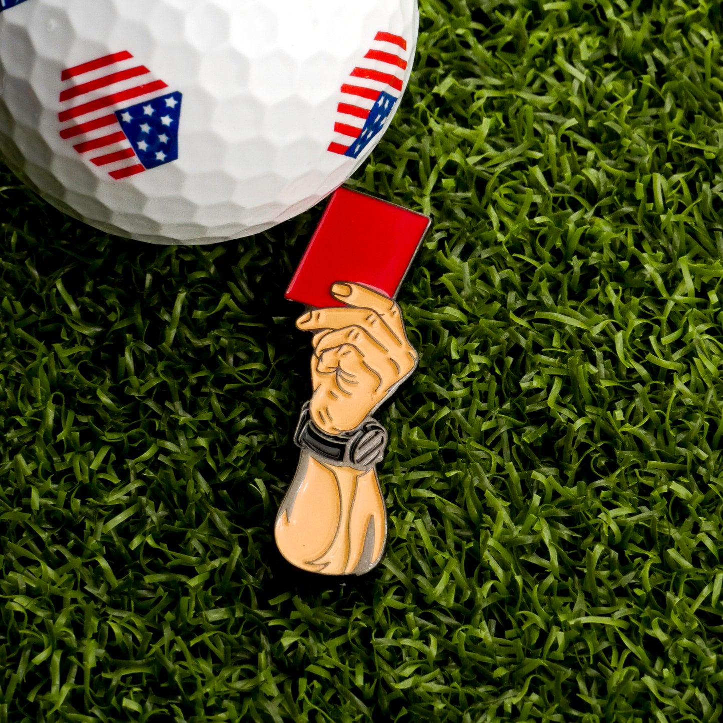 A close-up of an American flag golf ball with the Red Card Ball Marker by Ballmarkers.com, shaped like a referee's hand, displayed on green artificial turf.