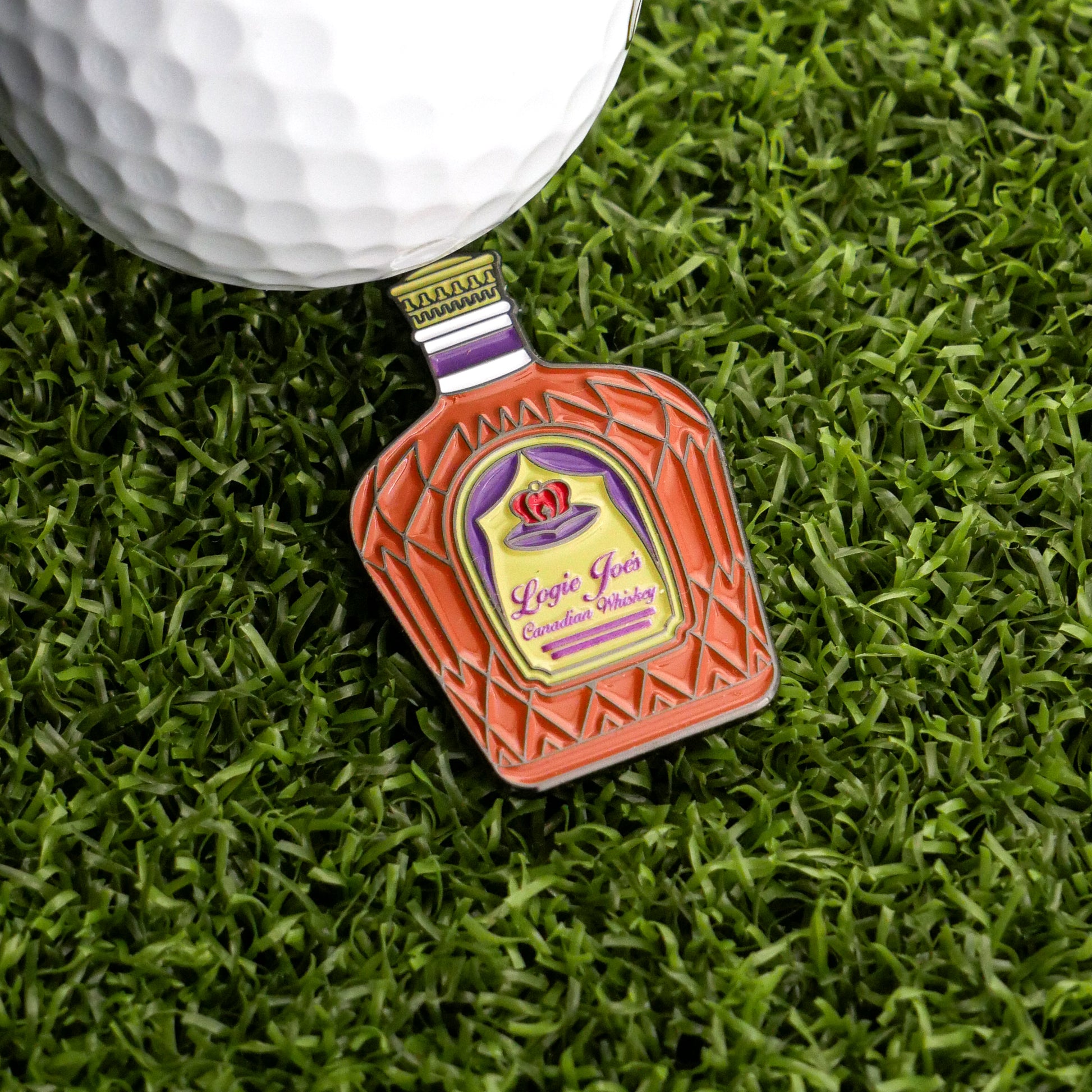 A bottle-shaped Canadian Whiskey Ball Marker by Logie Joe's Golf, featuring a colorful label, rests on green grass beside a white golf ball—making for a standout and unique golf accessory.
