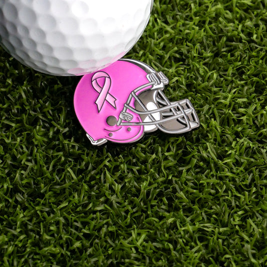 A golf ball sits on artificial grass near the Ballmarkers.com Football Helmet - Breast Cancer Ball Marker, featuring a pink ribbon—ideal for golfers supporting breast cancer awareness on and off the course.